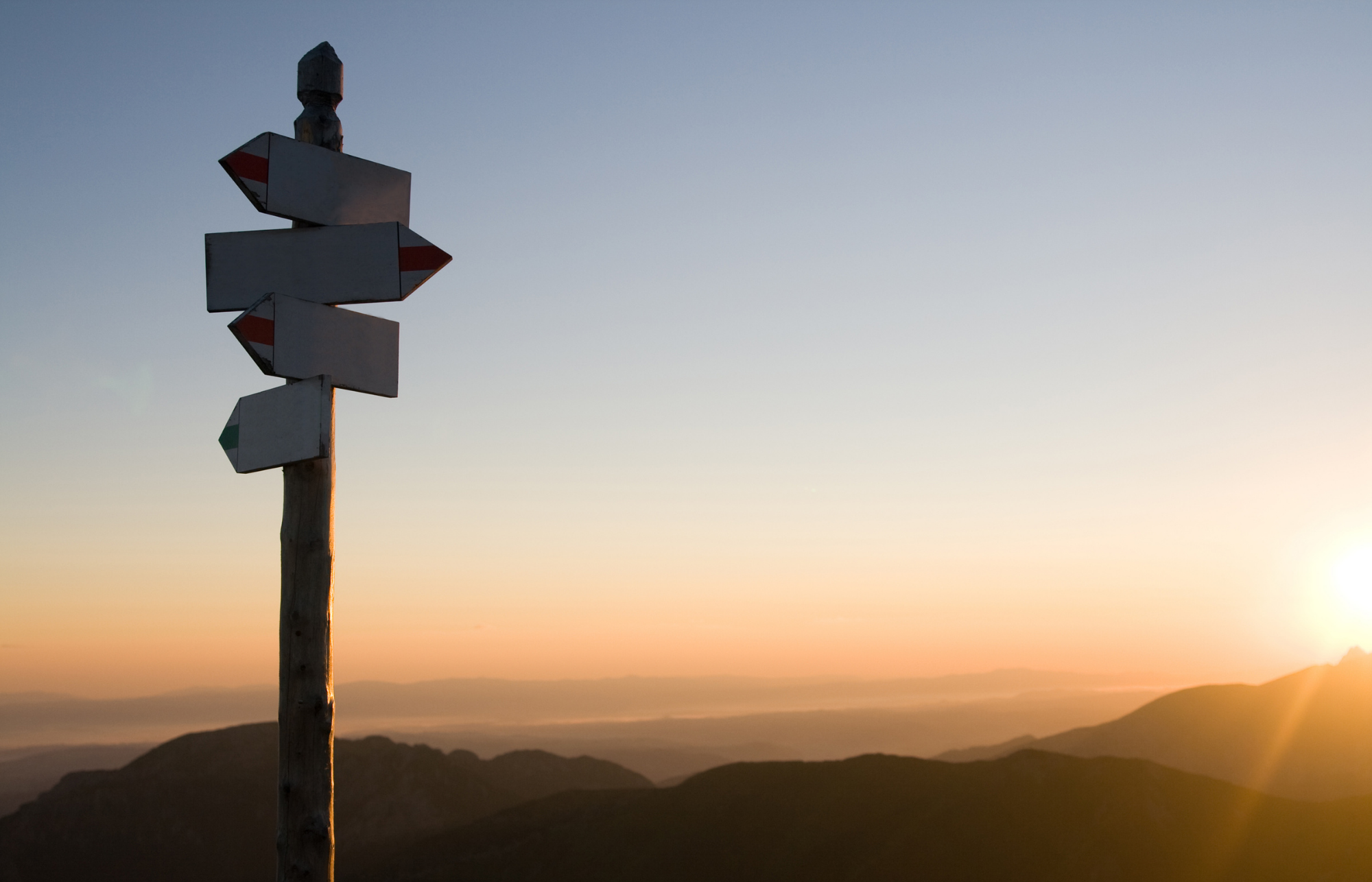 17 A sign post with arrows pointing in different directions in front of a landscape of mountains and a sunrise.